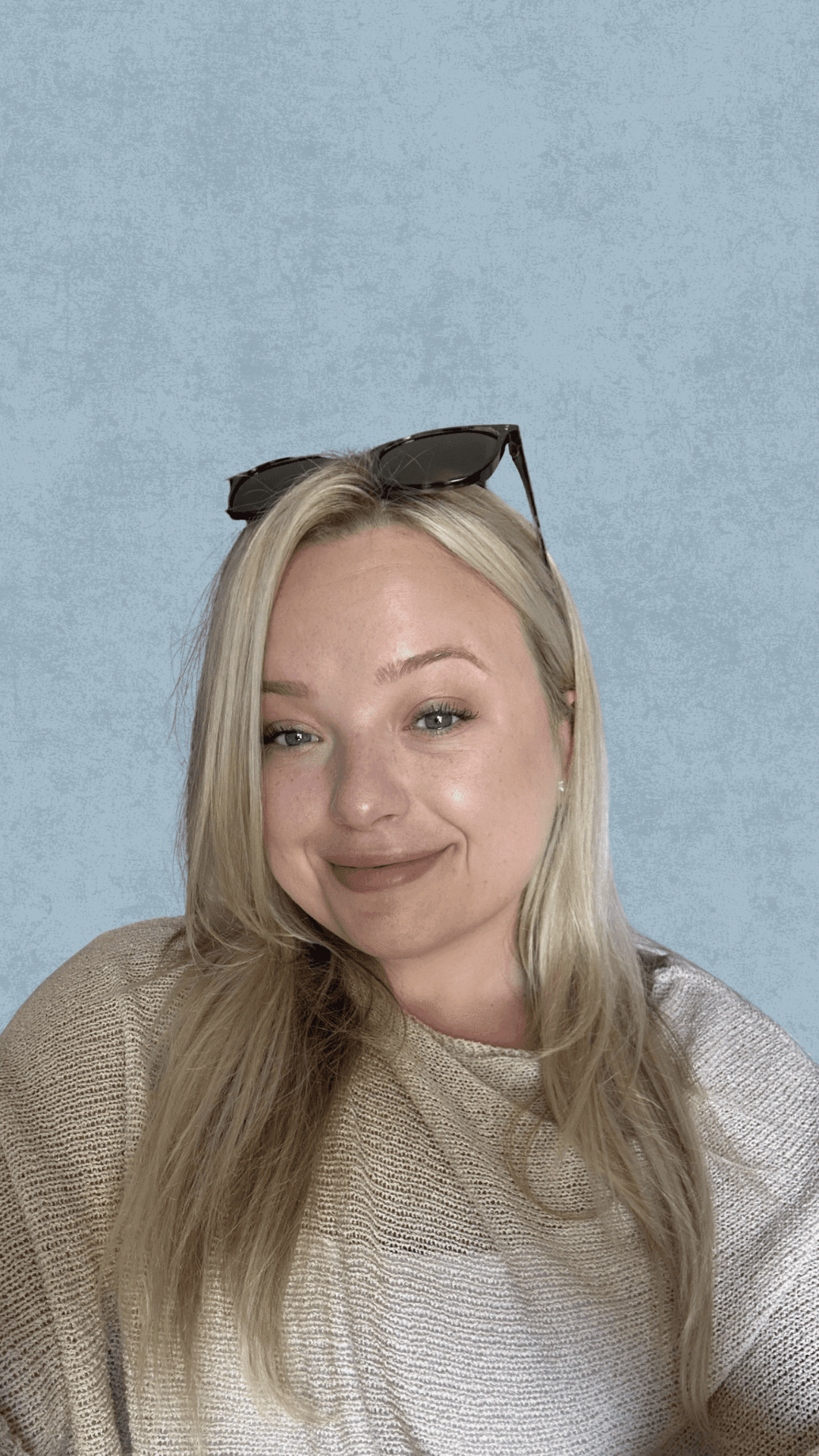 Headshot of a smiling blonde person wearing a beige sweater and sunglasses on their head, against a light blue textured background.