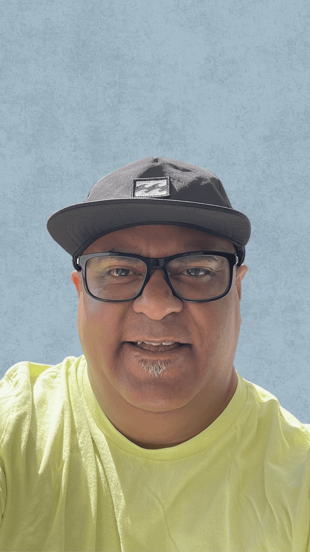 Headshot of a person wearing glasses, a black baseball cap and a bright yellow shirt, against a light blue textured background.